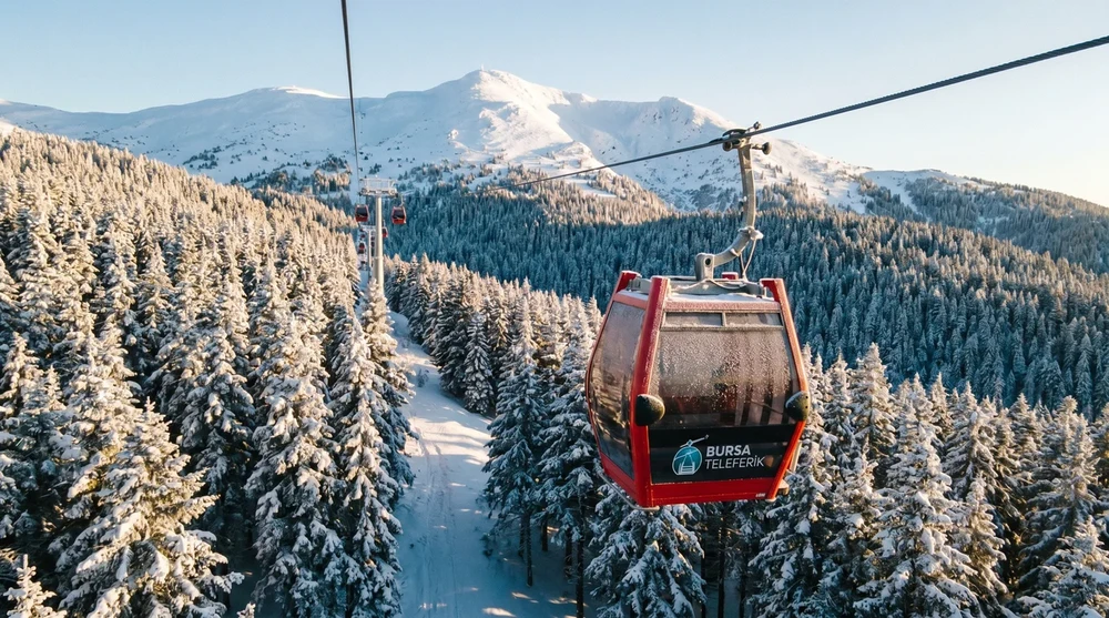 Red Bursa Teleferik cable car cabin over snow-covered pine forest with mountain peaks visible