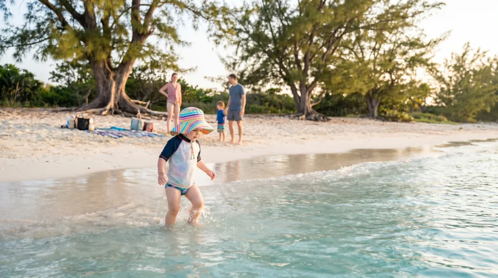 Toddler wading in ankle-deep clear water at Rum Point beach in Grand Cayman with pine tree shade