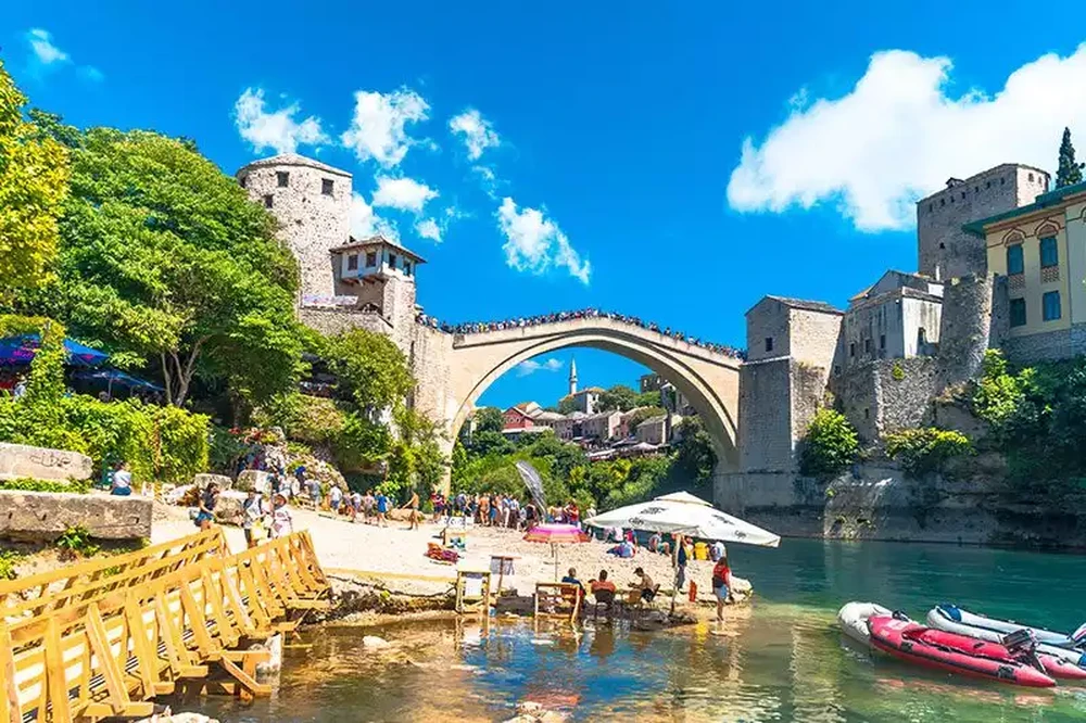 River-level view of Stari Most stone arch with tourists on the riverbank and boats in the foreground
