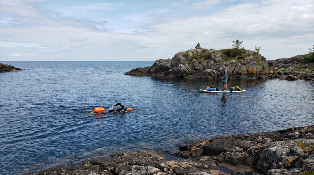 Swimmer and paddleboarder crossing a clear channel toward a small rugged islet away from the crowds