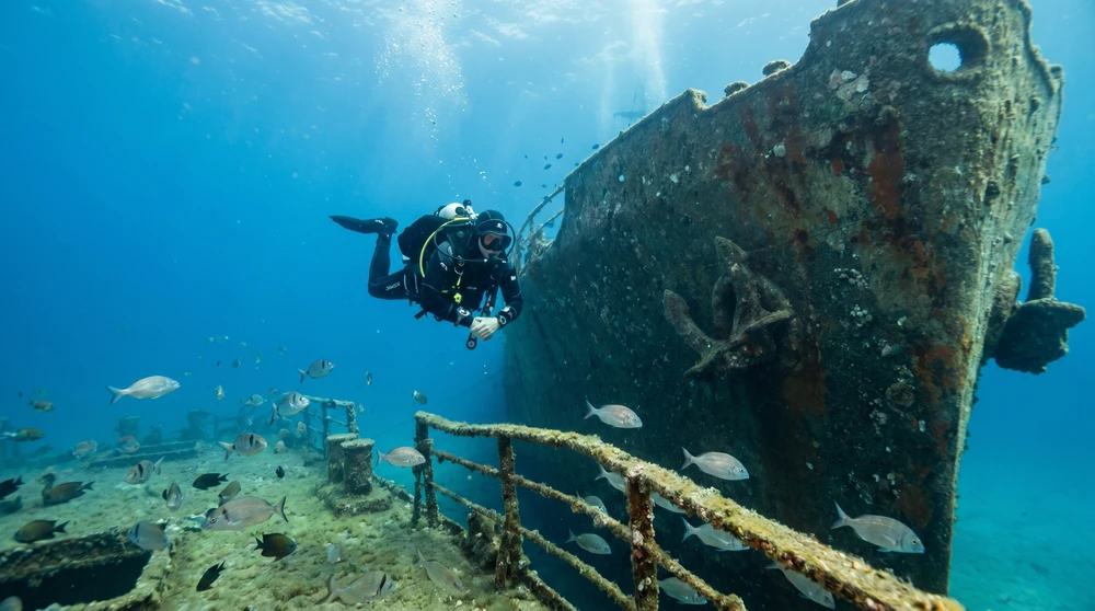 Scuba diver descending alongside a large sunken shipwreck in clear blue Mediterranean water