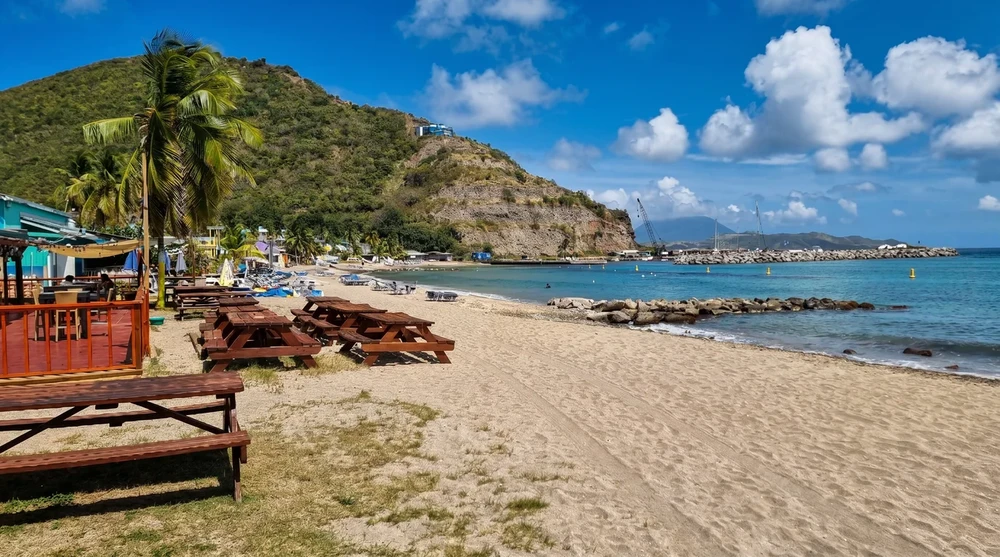South Frigate Bay shoreline with wooden picnic tables, beach bar deck, rocky breakwater and the long calm Caribbean curve under blue sky.