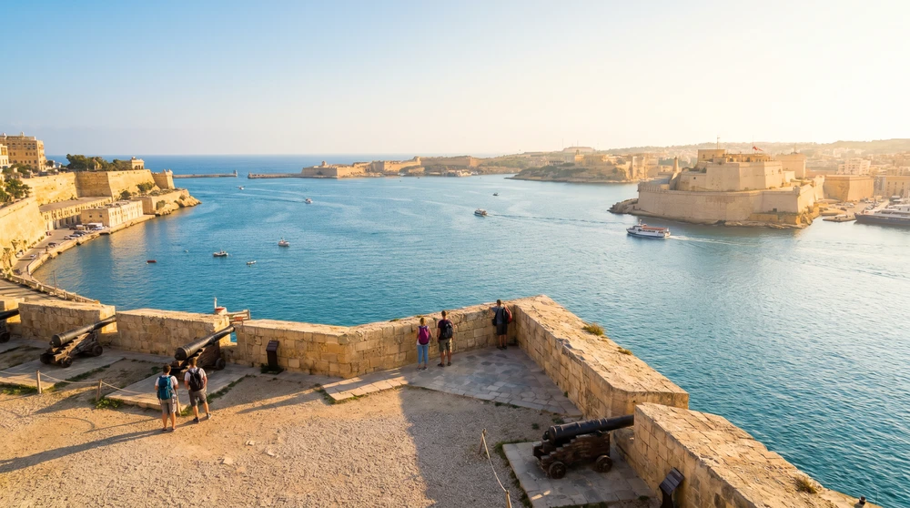 Panoramic view across the sunlit Grand Harbour toward golden fortifications from a peninsula viewpoint