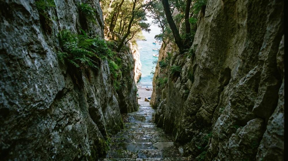 Carved stone steps descending a narrow limestone gorge toward a secluded turquoise pebble cove