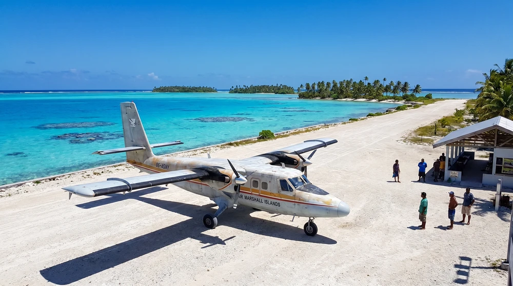 A small Air Marshall Islands aircraft parked on a remote atoll airstrip with a turquoise lagoon stretching into the background under a clear sky. These small planes are the only way to reach the outer atolls.