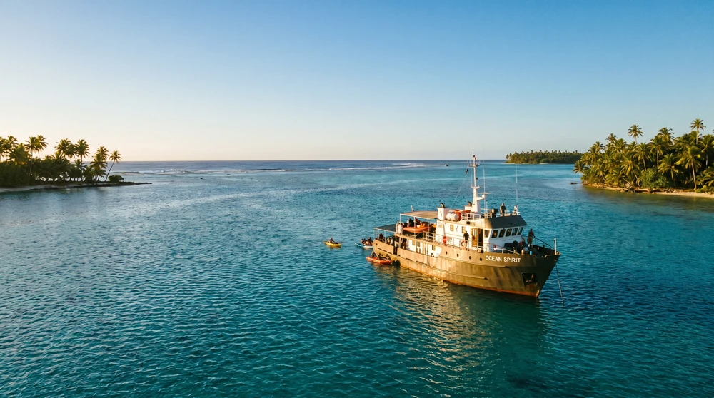 A calm Pacific lagoon under clear dry-season skies with an expedition ship anchored offshore in the Marshall Islands