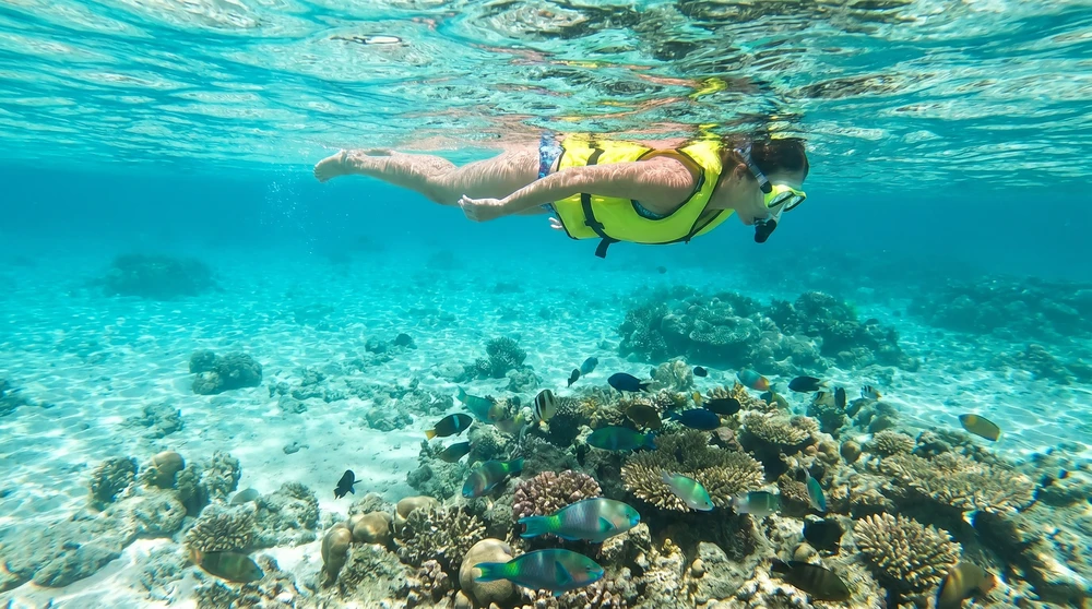 Nichtschwimmer mit gelbem Schnorchelgurt schwebt entspannt über einem flachen Korallenriff in einer Lagune der Marshall-Inseln, kristallklares Wasser mit Rifffischen darunter.