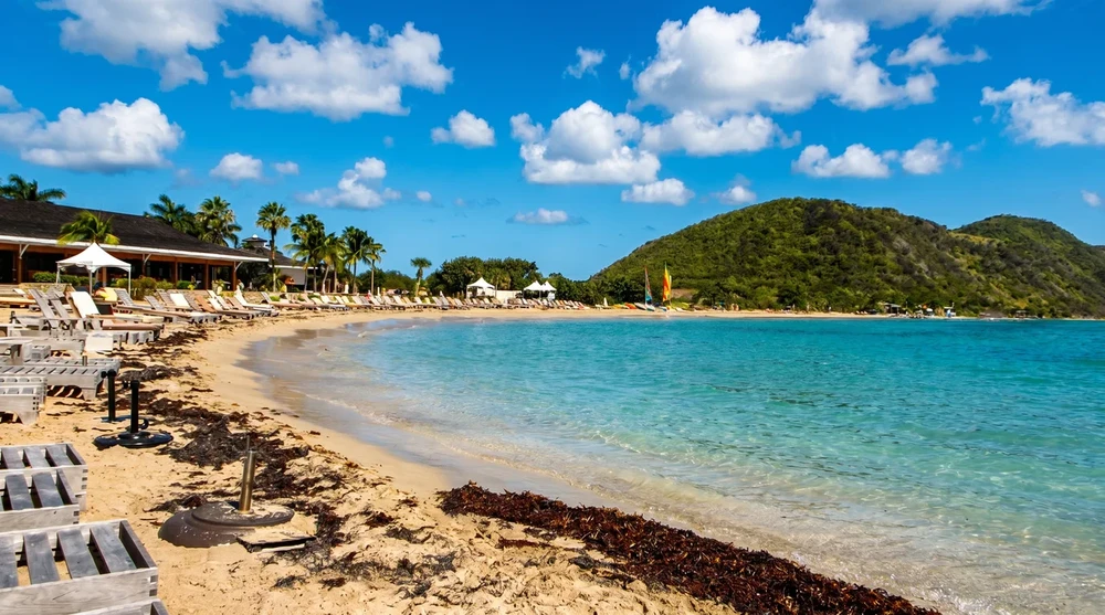 South Friars Bay long sandy beach with empty wooden lounge chairs, light seaweed line, turquoise calm water and a low-rise beachfront resort tucked into palms.
