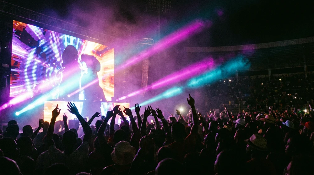 Anonymous crowd hands raised in silhouette against a magenta and cyan carnival stage at night.