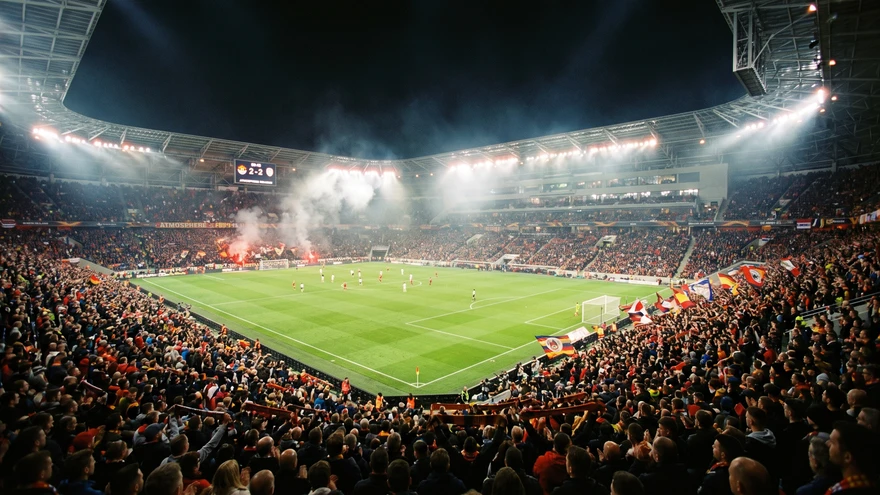 Packed football stadium at night under floodlights with colorful fans in the stands