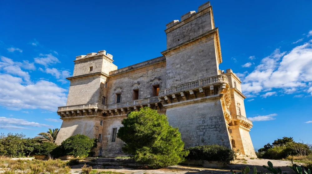 A large fortified coastal tower in Malta with a balustraded gallery seen from below against a blue sky