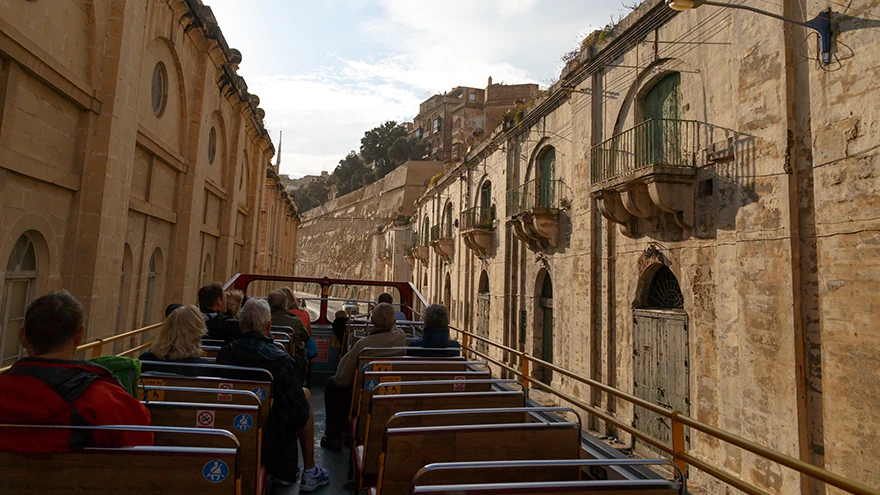 Fahrgäste auf dem offenen Oberdeck eines Hop-on-Hop-off-Sightseeing-Busses in einer historischen Steinstraße in Malta
