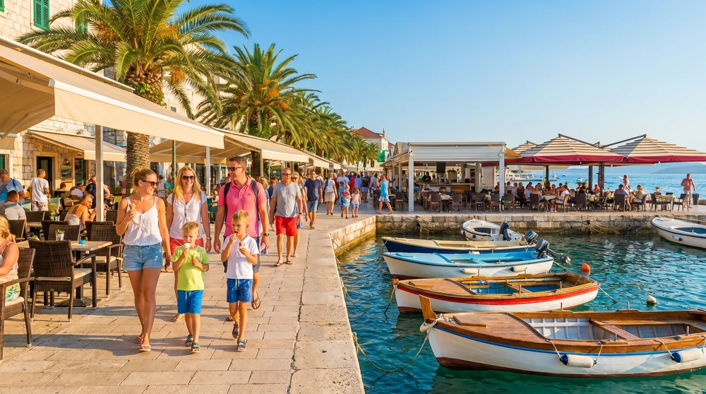 Families strolling a relaxed seaside promenade with boats and waterfront restaurants on a summer afternoon