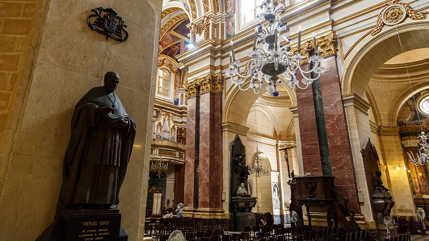 Innenraum der Paulskathedrale Mdina mit einer bronzenen Erzbischofsstatue, Marmorsäulen und einem hängenden Kristallleuchter