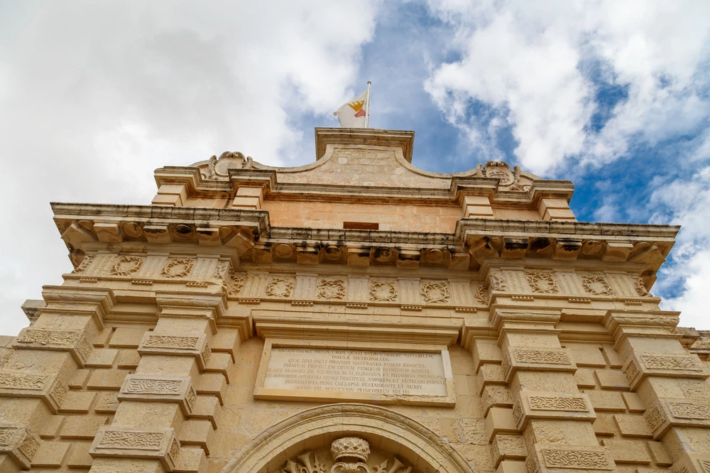Looking up at Mdina's Baroque Main Gate with the Maltese flag and carved Latin inscription, Malta
