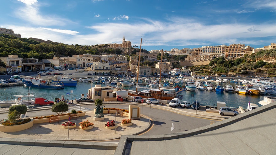 Panoramic view of Mgarr Harbour in Gozo with moored fishing boats, the marina and Our Lady of Lourdes church on the hillside, Malta