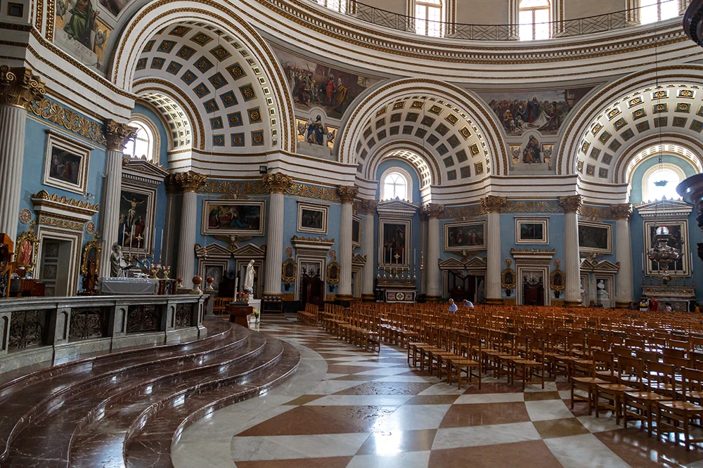 Das weitläufige, mit Stühlen bestückte Kirchenschiff und das blau gewölbte Innere der Basilika von Mosta, Malta