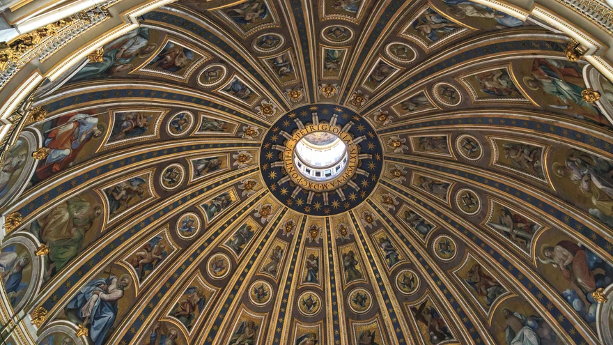 Upward view of the Michelangelo dome interior at St. Peter's Basilica showing the central oculus, gold mosaic saints in circular panels, and the Latin inscription band