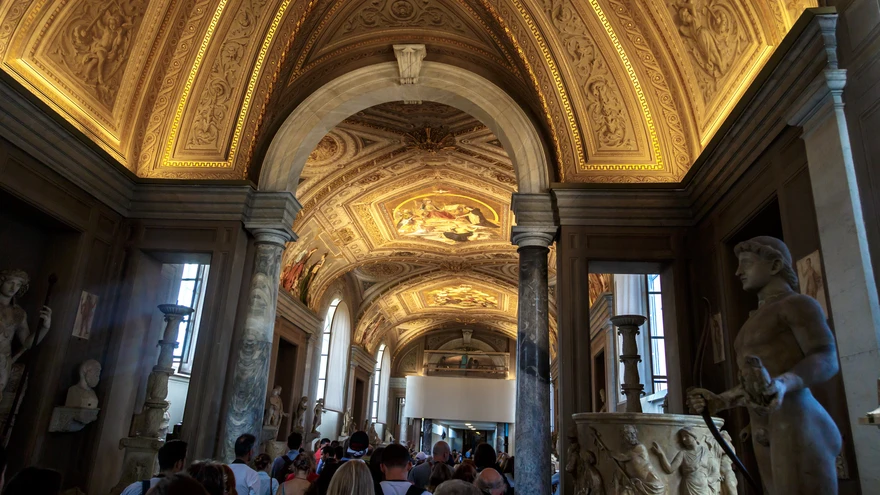 Crowded corridor of the Galleria dei Candelabri in the Vatican Museums, with visitors moving beneath a spectacular gilded barrel-vaulted ceiling and ancient Roman statues lining both sides