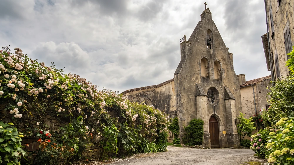 Rose-lined gravel path leading to the stone facade of Notre-Dame church in Castelmoron-d'Albret.
