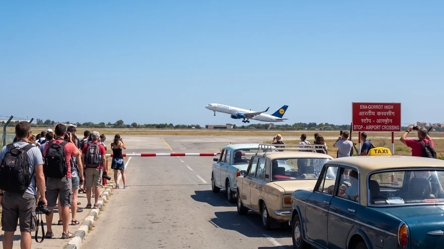 Pedestrians waiting behind a lowered barrier as a plane takes off from the runway ahead